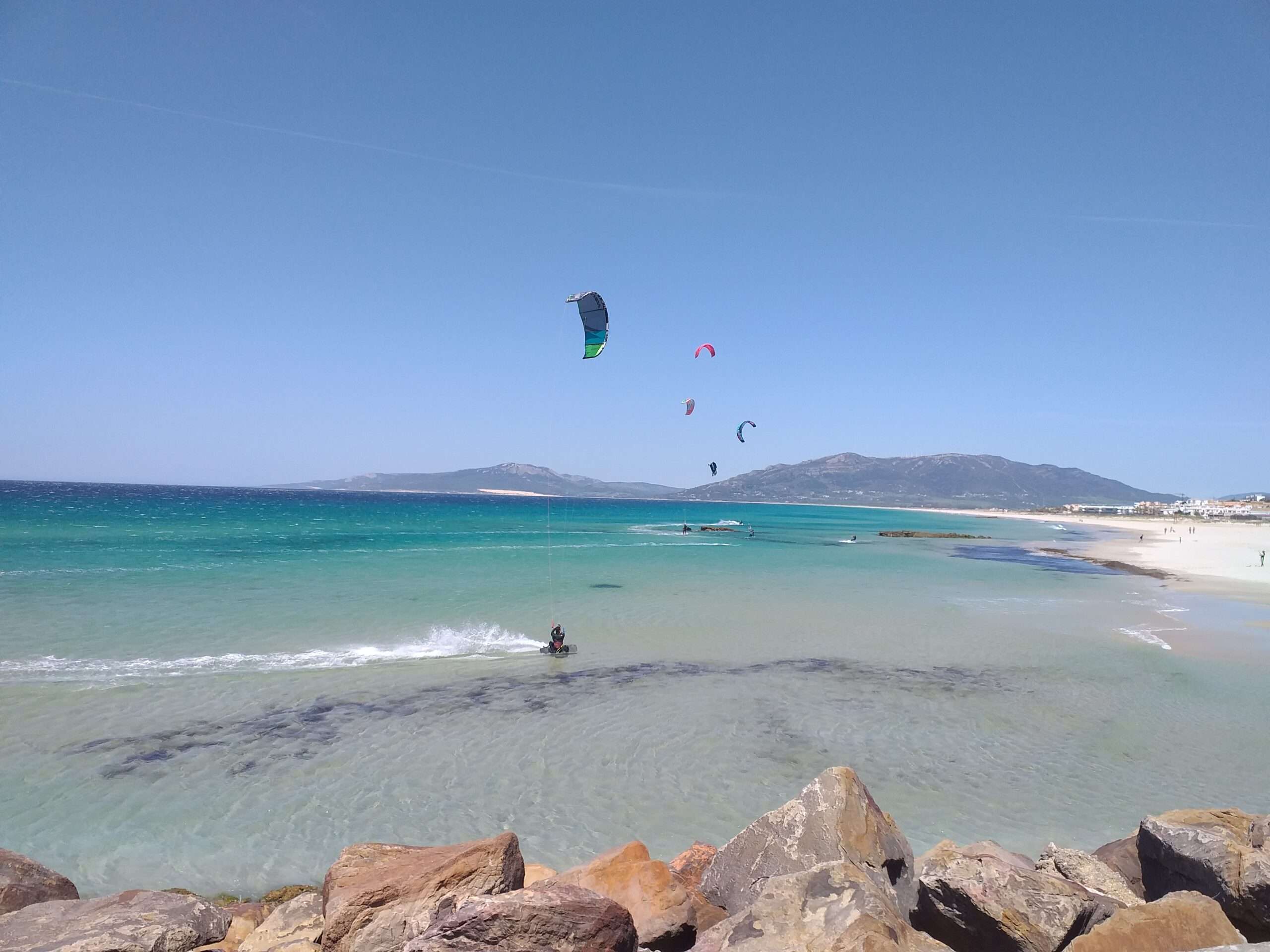 rocks, cristal blue water, white sandy beach, 5 people kitesurfing, mountains in the back