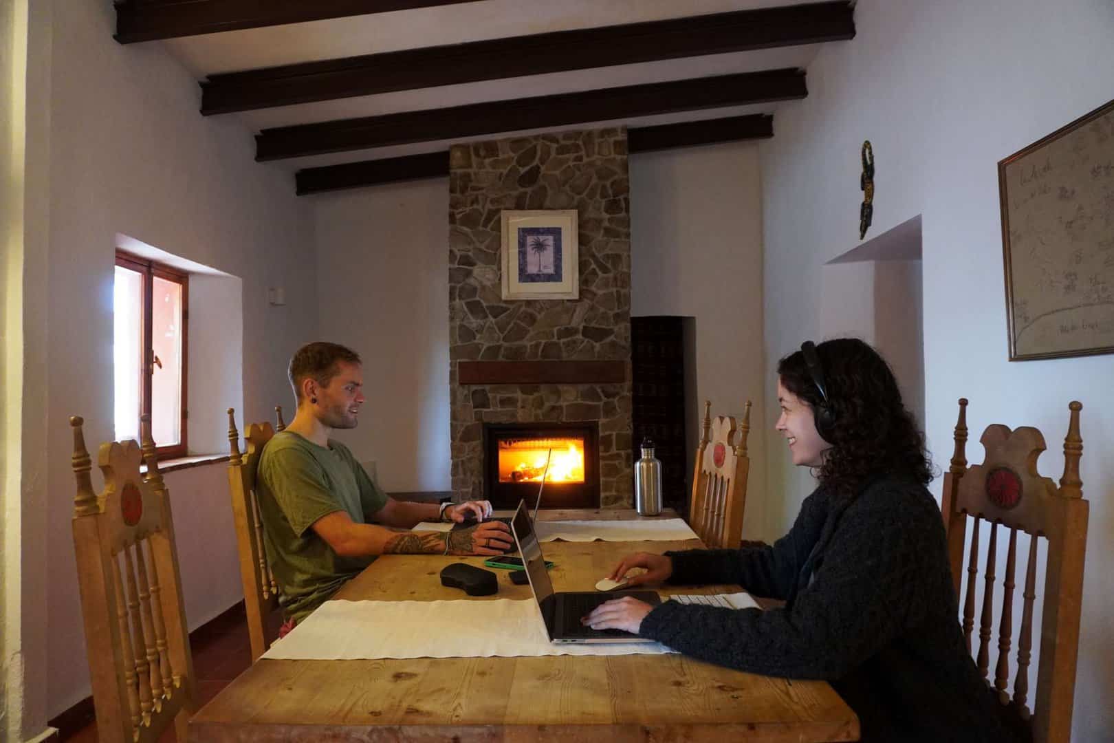 2 people sitting on a wooden table with their laptops, a fireplace in the background