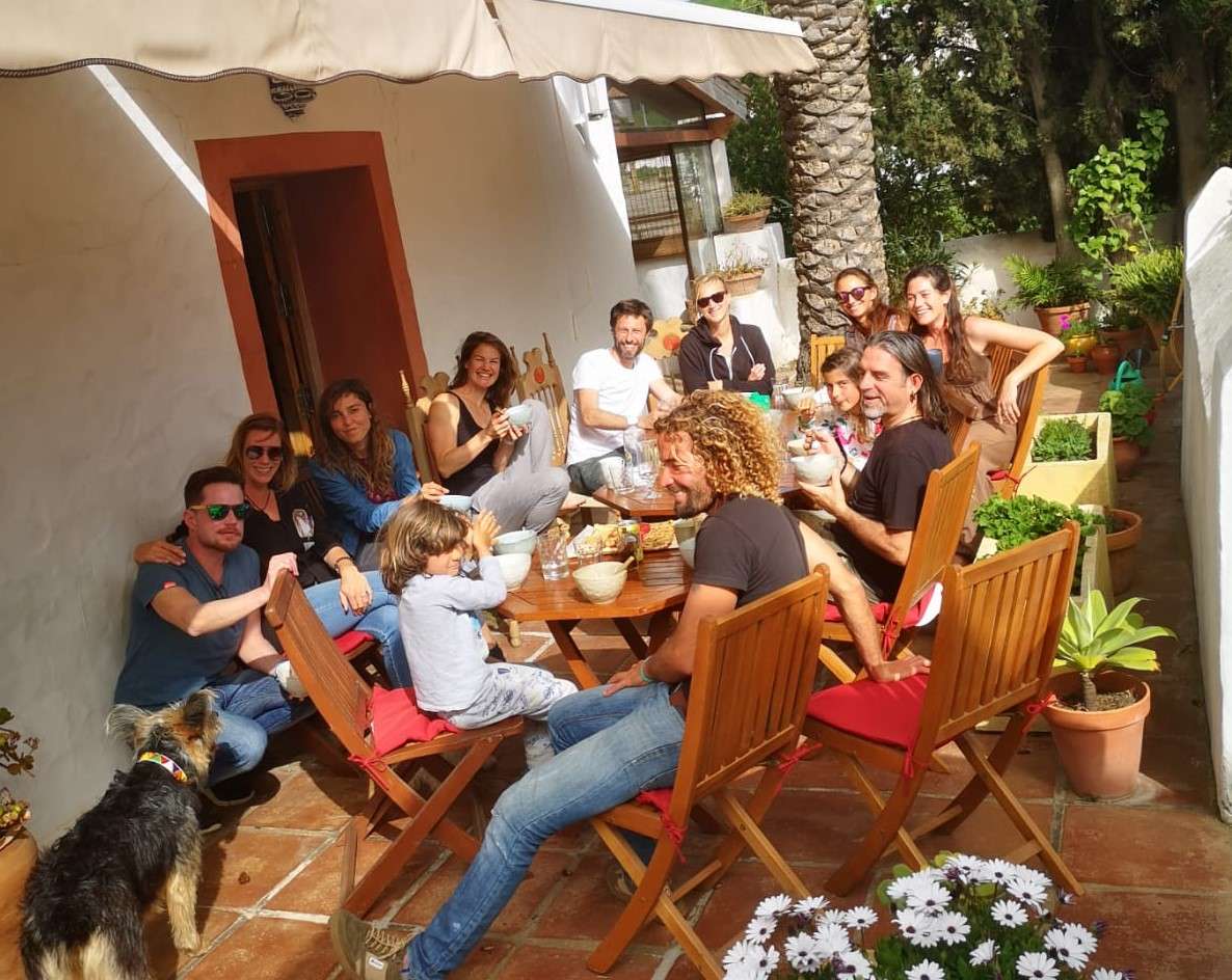 a group of 12 people, including 2 kids sitting on a big table in teh sun enjoying brunch, plants, flowers and a dog around them
