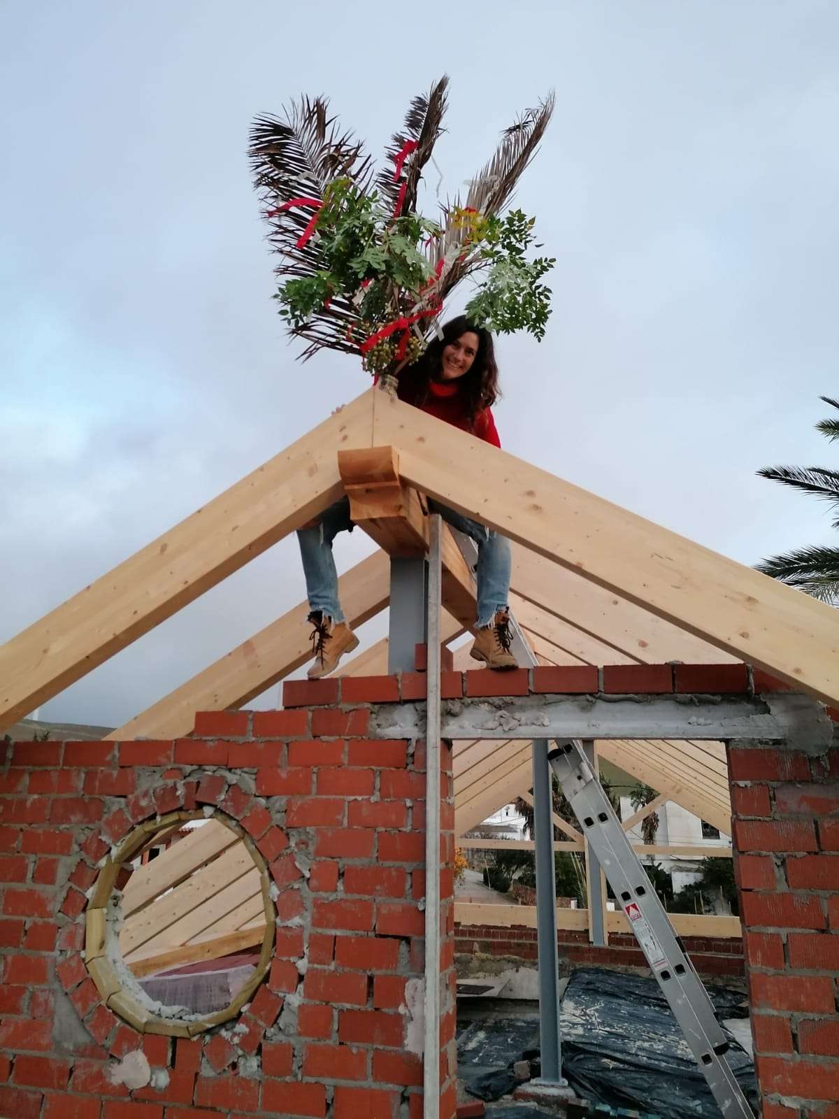 roof construction with a woman sitting on top of the wooden beam. The front of the roof is decorated with a bundle of palm tree leaves and coloured ribbon.