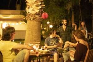 night time, a house in dthe background, a table with some people holding drinks, talking animatedly in teh front, decoration of lights and balloons around them in the garden and trees