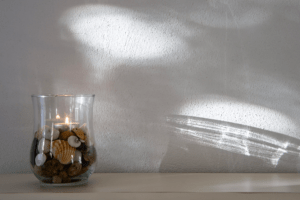 a jar with shells, stones and a lit candle agains a white wall with shadows
