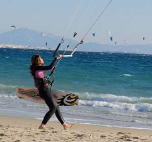 a girl flying a kite on the beach, board in hand, many other kites in the background on the water